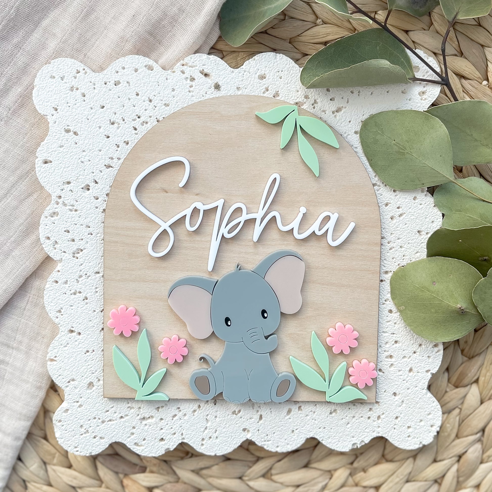 Decorative sign with an elephant and flowers on a textured surface with greenery.